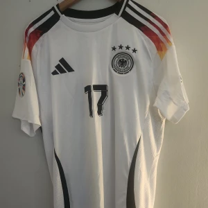 Tysklands Wirtz 17 fotbollströja Adidas - Snygg vit Tyskland fotbollströja från Adidas med Wirtz och nummer 17 på ryggen. Tröjan har svarta detaljer, färgglada axelpartier och officiella UEFA Foundation och EM 2024-patchar på ärmarna. Klassisk kortärmad modell i lätt funktionsmaterial. Storlek XL. Tröjan är inte äkta!
