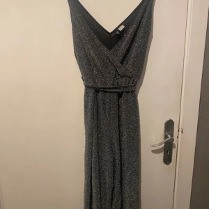 Glittrig svart jumpsuit med omlott - Snygg svart jumpsuit med glittrigt tyg och omlott-ringning. Vida ben och knytband i midjan ger en chill och trendig look. Perfekt för dig som vill sticka ut med lite extra glitter. Materialet är mjukt och följsamt, passar dig som gillar bekväma men stiliga plagg.