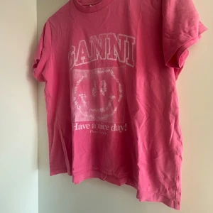 Rosa Ganni t-shirt med smileytryck - Cool rosa t-shirt från Ganni med stort vitt tryck framtill, där det står 'GANNI' och 'Have a nice day!' tillsammans med en smiley. Klassisk rund hals och korta ärmar. Skön bomullskvalitet som känns mjuk mot huden. Perfekt för en avslappnad och trendig look.