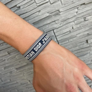 Blått Christian Dior J'Adior armband - Snyggt blått tygarmband inspirerat av Christian Dior med texten 'J'Adior' broderad i vitt. Armbandet har justerbar knytning med två coola tofsar i ändarna. Perfekt accessoar för att lyfta din outfit med en exklusiv touch.