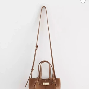 Brun axelväska från Lindex med gulddetalj - Snygg brun axelväska från Lindex som är slutsåld med justerbar lång rem och två handtag. Perfekt storlek för det viktigaste. Har fortfarande kvar kvitto samt plasten över då den är helt ny och aldrig kommit till användning💗