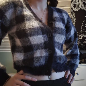 Rutig fluffig kofta i svart och grått - Supermjuk och fluffig kofta med stora svarta och grå rutor. Croppad modell med v-ringning och stora knappar framtill. Perfekt för dig som gillar mysiga vibes och vill sticka ut lite extra. Långärmad och riktigt cozy känsla.