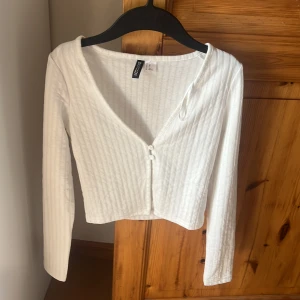 Vit ribbad topp från H&M DIVIDED - Säljer en vit ribbad topp från H&M DIVIDED i storlek XS. Toppen har v-ringning, lång ärm och två knappar framtill. Croppad modell som är perfekt att styla med höga jeans eller kjol. Enkel och stilren design med smala vertikala ränder.