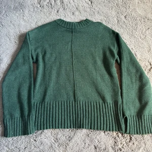 Grön stickad tröja från Vila - Mysig grön stickad tröja från Vila i storlek S. Tröjan har ribbade muddar och bred ribbad nederkant, samt en snygg söm mitt på ryggen. Perfekt för lager-på-lager och chill dagar.