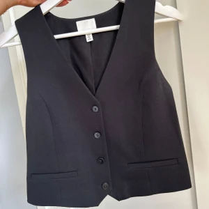 Svart kostymväst från H&M - Klassisk svart väst från H&M med snygg v-ringning och fyra knappar framtill. Använd ett fåtal gånger!