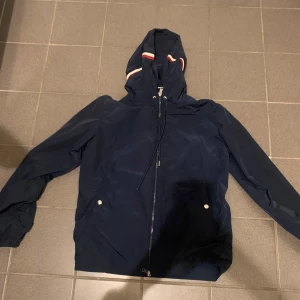 Mörkblå vindjacka från Moncler - Snygg mörkblå vindjacka från Moncler med huva och vita/röda detaljer. Jackan har dragkedja framtill, två fickor med tryckknappar och Moncler-logga på bröstet. Perfekt för blåsiga dagar och enkel att matcha med det mesta.
