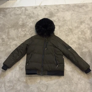 Grön pufferjacka Glorious Gangsta L - Snygg mörkgrön pufferjacka från Glorious Gangsta med svart fuskpäls på huvan och diskret mönster på ärmarna. Jackan har flera dragkedjefickor, ribbade muddar och en schysst oversized look. Perfekt för kalla dagar när du vill sticka ut lite extra.