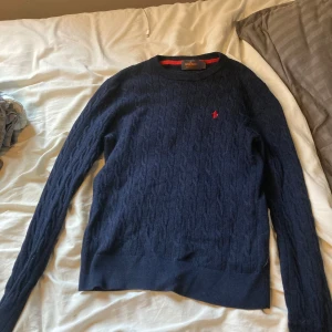 Mörkblå stickad tröja från Morris - Snygg mörkblå kabelstickad tröja från Morris i 100% merinoull. Tröjan har rund hals, långa ärmar och en röd broderad logga på bröstet. Perfekt för dig som gillar klassisk och stilren look med en twist. Använd få grejar pga krympt i tvätten s->xs