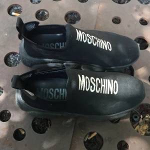 Svarta slip-on sneakers från Moschino - Svarta slip-on sneakers från Moschino med tydlig vit logga på ovansidan. Skorna har en rund tå, platt sula och är tillverkade i textilmaterial. Perfekta för en streetstyle-look och lätta att ta på tack vare elastisk öppning.