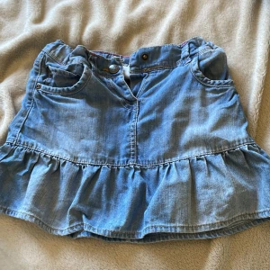 Blå jeanskjol med volang från FIX - Supersöt blå jeanskjol från FIX med volang nertill och broderad ficka bak. Är i storlek 116 men passar mig som brukar ha storlek 152 som minst och som störst S. Klassisk femficksmodell med knapp och dragkedja framtill. Snygga detaljer med rosa och röd randig insida i midjan och broderat H på bakfickan. 