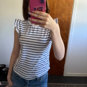 Randig volangtopp från SHEIN XS - Superfin topp från SHEIN i storlek XS med vita och blå ränder. Modellen är ärmlös med volangdetaljer på axlarna och rund halsringning. Perfekt för dig som gillar en söt och avslappnad stil. Tillverkad i mjuk bomull.