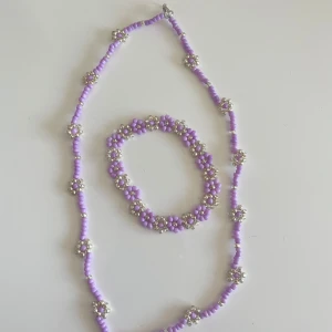 Lila pärlset halsband & armband - Supergulligt set med halsband och armband i lila och silverfärgade pärlor. Båda smyckena har små blommor av pärlor som ger en söt och lekfull vibe. Perfekt för att piffa upp din outfit med lite färg och charm.