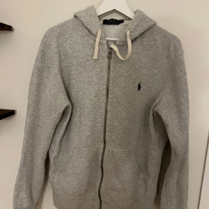Grå hoodie från Polo Ralph Lauren - Grå hoodie från Polo Ralph Lauren med dragkedja, huva med snörning och klassisk broderad logga på bröstet. Mjuk bomullskänsla, två fickor fram och ribbade muddar. Perfekt för chill dagar eller när du vill ha en clean look.