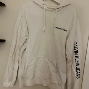 Vit hoodie från Calvin Klein Jeans - Vit hoodie från Calvin Klein Jeans med svart logga tryckt på bröstet och längs ena ärmen. Tröjan har huva med snörning och en stor magficka framtill. Perfekt för en clean och stilren look.