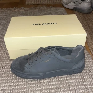 Mörkgrå sneakers från Axel Arigato - Snygga mörkgrå sneakers från Axel Arigato med stilren design. Skorna har ovandel i skinn och mocka, rund tå och platt sula. Guldtext med logga på sidan och innersulan. Perfekta för dig som gillar minimalistisk och clean stil. Storlek 44, aldrig använt helt nytt skick 