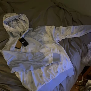 Ljusblå hoodie från HUGO BOSS med logomönster - Säljer en snygg ljusblå hoodie från BOSS med all-over broderat logomönster. Tröjan har huva, långa ärmar och ribbade muddar. Perfekt för dig som gillar stilrena och trendiga plagg med premiumkänsla. Aldrig använd förut pris lapp även kvar, storlek S