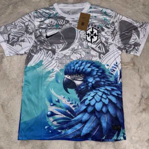 Brasilien x nike  fotbollströja  - Unik Nike fotbollströja för Brasilien (CBF) i Dri-Fit material. Tröjan är vit och grå med ett stort blått papegojmotiv framtill, coola grafiska detaljer och svart CBF-logga på bröstet. Kortärmad och perfekt för dig som vill sticka ut på planen med stil.