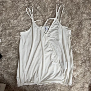 Vit omlott linnetopp från Amisu - Säljer en stilren vit linnetopp från Amisu med dubbla smala axelband och omlottdesign framtill. Toppen är ärmlös och har en mjuk, slät känsla i syntetmaterial. Perfekt för varma dagar och enkel att matcha med jeans eller kjol.