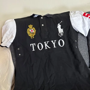 Svart och vit pikétröja Polo Ralph Lauren - Snygg svart pikétröja från Polo Ralph Lauren med vita ärmar och stora broderade detaljer. Framsidan har TOKYO-text, gult emblem och klassisk polospelare. Höger ärm har röd siffra 5. Perfekt för dig som gillar sportig och stilren look.