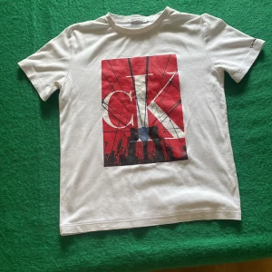 Vit Calvin Klein t-shirt med tryck - Vit t-shirt från Calvin Klein med stort grafiskt tryck i rött, svart och vitt på bröstet. Trycket har CK-logga och stadssilhuett. Klassisk passform och rund hals. Perfekt för dig som gillar streetstyle och snygga loggor.