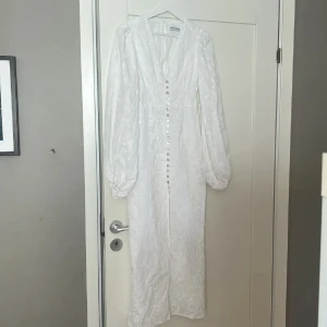 Vit långklänning med knappar från Adoore - Superfin vit långklänning från Adoore med v-ringning, långa puffärmar och dekorativa knappar framtill. Klänningen har ett subtilt blommönster i tyget och markerad midja för en elegant siluett. Perfekt för speciella tillfällen!