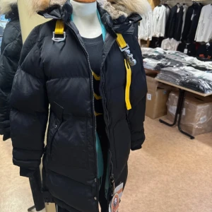 Svart Parajumpers dunjacka med päls - Svart dunjacka från Parajumpers med gul detalj vid dragkedjan och stor huva med fluffig päls. Jackan har patch på ärmen, flera fickor och coola spännen. Perfekt för kalla dagar när du vill hålla stilen och värmen.