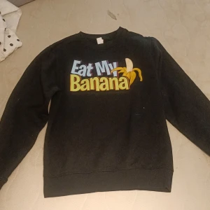 Svart sweatshirt med banan-tryck - Svart sweatshirt med färgglatt tryck där det står 'Eat My Banana' och en tecknad banan. Tröjan har rund hals och långa ärmar, perfekt för dig som gillar humoristiska plagg och vill sticka ut lite extra.