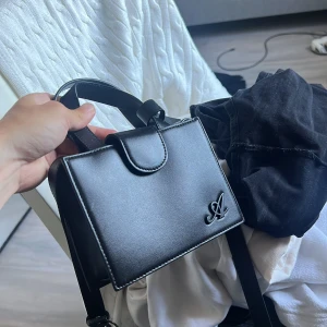 Axel arigato väska svart - Snygg svart axel arigatoväska i stilren, boxig modell med lock och magnetstängning. Väskan har ett elegant A-emblem i metall på framsidan och justerbar axelrem. Perfekt storlek för det viktigaste och passar till många outfits.