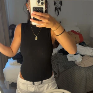 Svart ärmlös body med öppen rygg - Säljer en svart, tight och ärmlös body med hög hals och en riktigt snygg öppen rygg. Perfekt för dig som vill sticka ut lite extra! Toppens material känns stretchigt och följsamt, vilket gör den superbekväm att bära hela dagen.