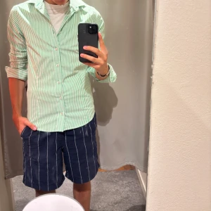 Grön- och vitrandig skjorta - Snygg skjorta med smala gröna och vita ränder, klassisk krage och knäppning framtill. Skjortan har lång ärm och en slim passform som sitter tajt men bekvämt. Perfekt för dig som gillar en fräsch och stilren look.