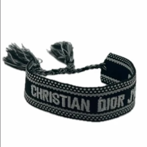 Christian Dior armband - Svart och silverfärgat tygarmband från Christian Dior med texten 'J'Adior'. Armbandet har flätad knytning och tofsar i ändarna. Snygg detalj med broderade kanter och kontrasterande färger som ger en lyxig och trendig vibe.