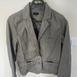 Grå kavaj från Lindex, stl 38 - Snygg grå kavaj från Lindex i storlek 38. Klassisk modell med två knappar framtill, tre fickor och markerad krage. Tillverkad i bomull för en skön känsla och passar perfekt till en chill eller dressad look.