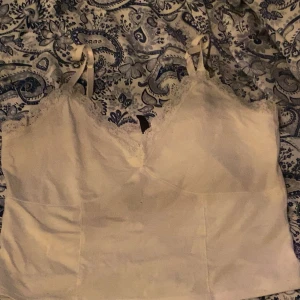 Vit spetslinnetopp från H&M Divided - Supersöt vit linnetopp från H&M Divided med tunna axelband och spetsdetaljer längs v-ringningen. Toppen har en mjuk, slät yta och är perfekt att styla med jeans eller kjol. Enkel och fräsch look med romantisk touch. Strl L men skulle säga M eller stor S