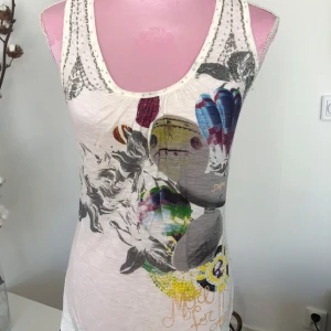 Desigual linnetopp med färgglatt tryck - Unik linnetopp från Desigual i vitt med färgglada tryck, ballonger och textdetaljer. Toppen har broderad logga på ryggen, knytning i ryggen och coola detaljer framtill. Perfekt för dig som gillar statement-plagg och kreativ design.