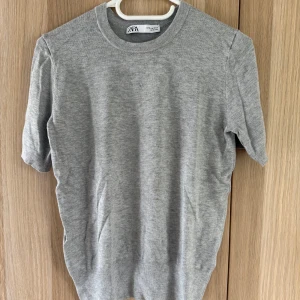 Grå kortärmad tröja från Zara - Enkel och stilren grå tröja från Zara i mjukt material. Tröjan har rund halsringning och korta ärmar, perfekt för en clean och avslappnad look. Passar till allt och är lätt att matcha med andra plagg.