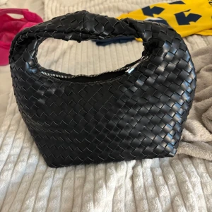 Svart flätad axelväska i skinn - Säljer en svart axelväska med snygg flätad design och dragkedja upptill. Väskan har en mjuk, rundad form och ett rejält handtag som gör den enkel att bära. Perfekt för dig som gillar stilrena accessoarer med lite edge.