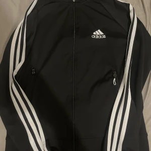 Svart Adidas vindjacka med vita ränder - Svart vindjacka från Adidas med klassiska vita ränder längs ärmarna och vit logga på bröstet. Jackan har hel dragkedja framtill och två fickor med dragkedja. Perfekt för en sportig och avslappnad stil.