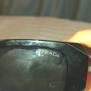 Svarta solglasögon från Prada - Snygga svarta solglasögon från Prada med rektangulära bågar och breda skalmar. Klassisk och stilren design med mörka glas och diskret Prada-logga på linsen. Perfekta för dig som vill ha en clean och lyxig look.