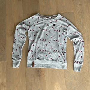 Grå tröja med fågelmönster Naketano M - Grå långärmad tröja från Naketano i storlek M, dekorerad med små röda och svarta fåglar över hela tyget. Tröjan har rund halsringning, ribbade muddar och en snygg brun detalj vid nederkanten. Perfekt för dig som gillar unika prints.