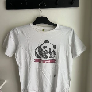 Vit panda t-shirt WWF 11-12 år - Vit t-shirt från WWF med en stor svart panda-illustration och texten 'EST. 1961' i rosa band på bröstet. Klassisk rund hals och korta ärmar. Perfekt för dig som gillar djur och vill ha en chill och enkel stil.