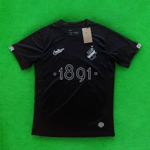 Svart AIK fotbollströja Nike 1891 - AIK fotbollströja i svart från Nike med trycket '1891' på bröstet, AIK:s klubbmärke och retro Nike-logga. Tröjan är kortärmad och tillverkad i ett lätt, sportigt material som andas. Perfekt för dig som vill visa ditt AIK-stöd på match eller träning.
