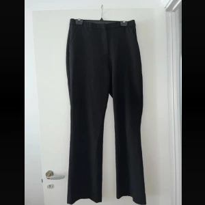Svarta kostymbyxor - Klassiska svarta kostymbyxor med raka, lite vidare ben och diskreta fickor bak. Byxorna har dragkedja och hake i midjan. Snyggt skräddad look som passar till många olika stilar. Materialet känns mjukt och har en lätt glans. Sliten nertill se sista bild. 