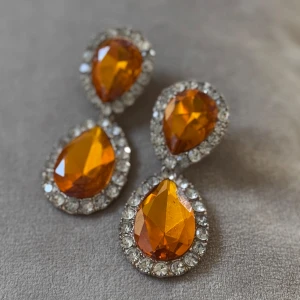Droppformade statementörhängen i orange och guld - Två par droppformade statementörhängen. Ena paret har stora orange stenar omgivna av glittriga stenar i silver, medan det andra paret har guldiga ramar med spegelblanka inslag. Perfekta för att sticka ut och ge din look extra edge.
