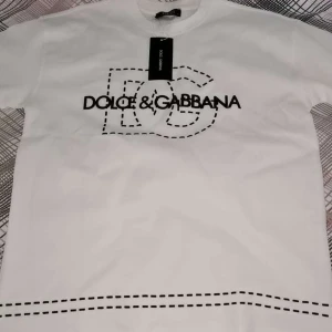 Vit Dolce & Gabbana t-shirt - Vit t-shirt från Dolce & Gabbana med svart tryck och coola broderade detaljer. Klassisk passform med korta ärmar och rund halsringning. Stora DG-bokstäver och logga framtill ger en stilren och trendig look. Perfekt för dig som gillar exklusiva märken.