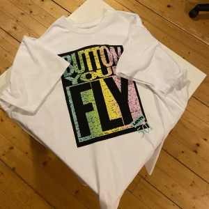 Vit t-shirt Levi's Button Your Fly - Vit t-shirt från Levi's med stort färgglatt tryck på framsidan där det står 'Button Your Fly'. Trycket har pastelliga toner i grönt, gult och rosa. Klassisk passform med rund hals och korta ärmar. Perfekt statement-plagg för dig som gillar retrostil.