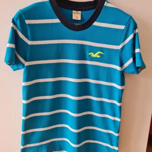 Blå och vit randig t-shirt från Hollister strl S - Snygg blå t-shirt från Hollister med vita ränder och korta ärmar. Den har en mörkblå krage och ett litet gult logotryck. Storlek S. Kan hämtas i Vallentuna.