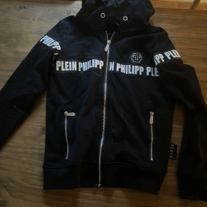Svart zip hoodie från Philipp Plein  - Svart hoodie med dragkedja från Philipp Plein. Stor logga och text på både fram- och baksida samt ärmar. Två fickor med dragkedja framtill.
