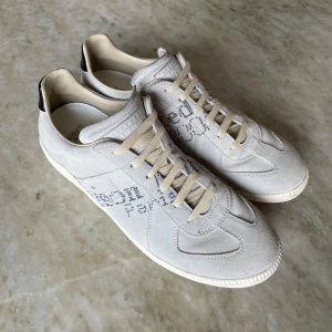 Vita sneakers från Maison Margiela - Maison Margiela sneakers i vitt mocka med limited edition textdetalj på sidan och svart läderhäl. Klassisk rund tå och snörning. Tillverkade i Italien, stilrena och minimalistiska med en lyxig känsla. Limited edition köpta nya på vestiere collective, därför garanteras äkthet, använda tre gånger