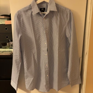 Blåvit randig skjorta från H&M - Klassisk blåvit randig skjorta från H&M i slim fit-modell. Skjortan har långa ärmar, knappar framtill och är tillverkad i en mjuk blandning av polyester och bomull. Perfekt för en stilren och fräsch look.
