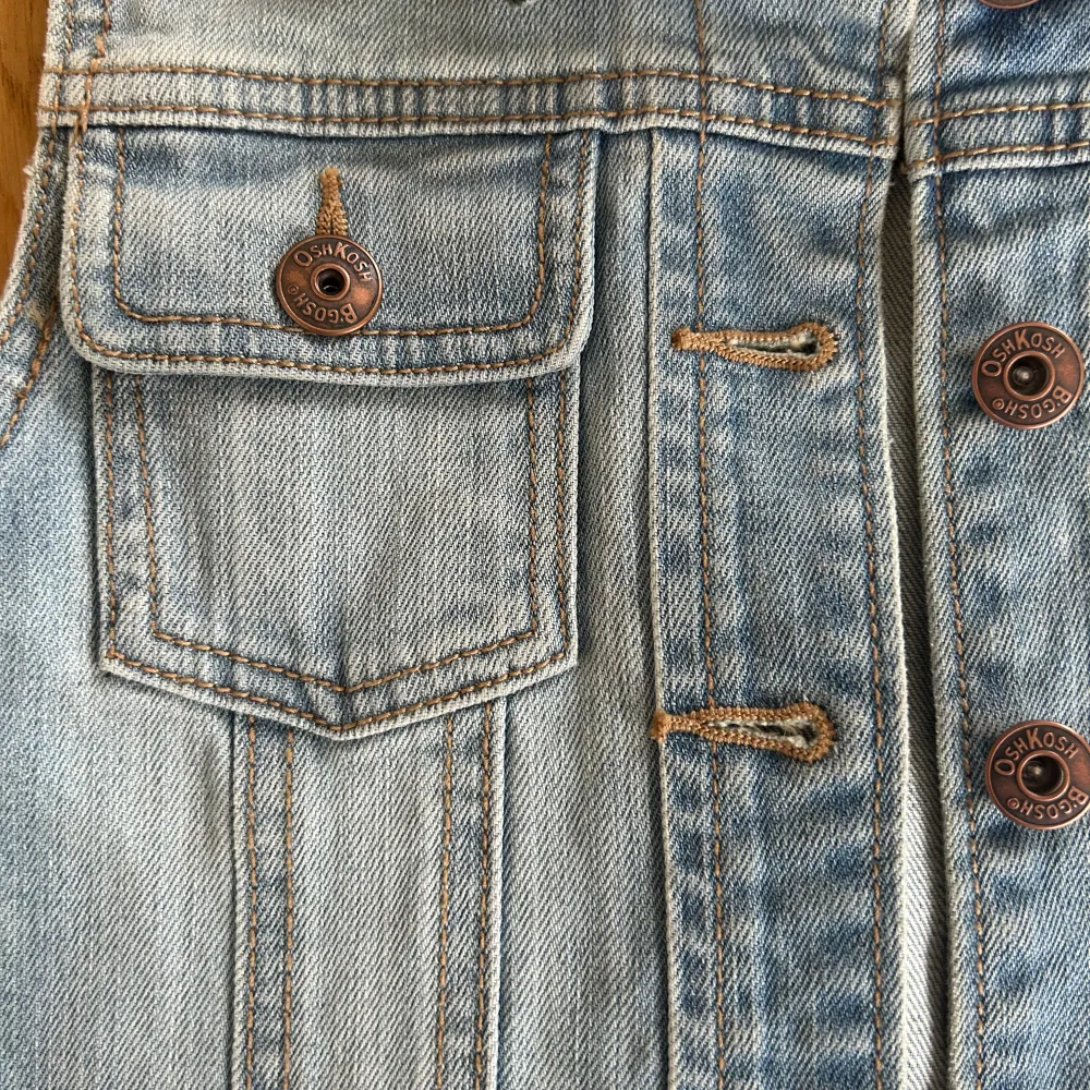 Säljer en klassisk ljusblå jeansväst från OshKosh B’gosh med bruna knappar och två bröstfickor med lock. Storlek barn 8 år.. Takit.